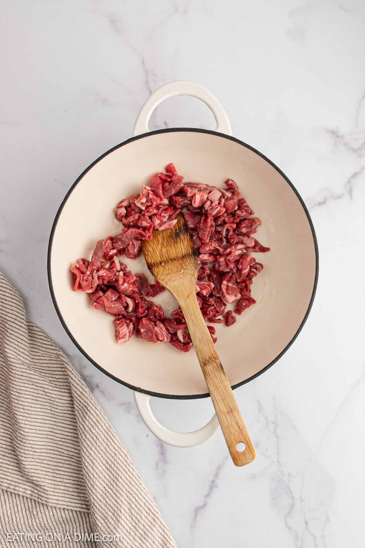 A white pot with sliced raw beef and a wooden spatula inside, ready to start beef fried rice, sits on a marble countertop next to a beige-striped towel.
