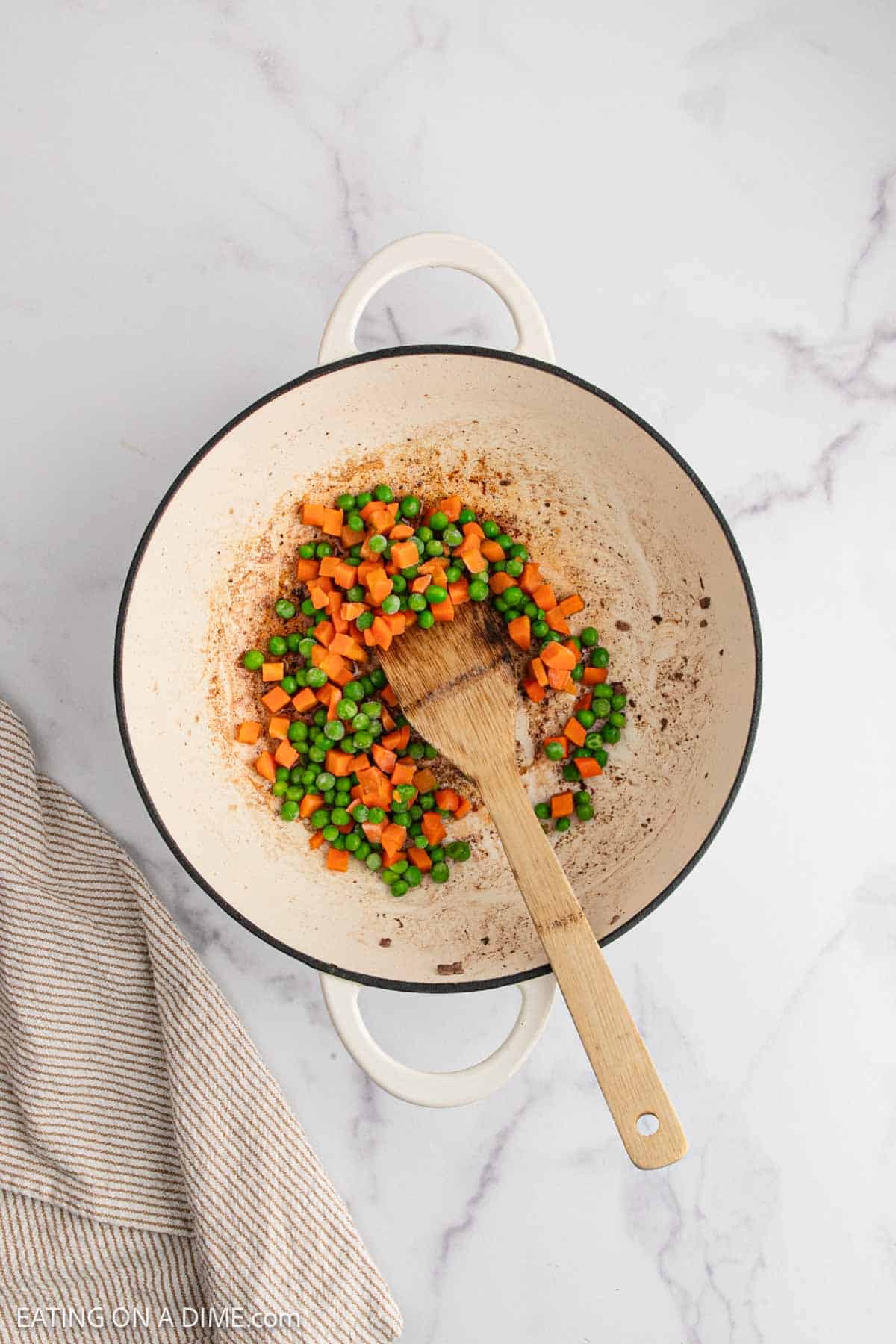 A white pot on a marble surface contains diced carrots and green peas being stirred with a wooden spoon—perfect veggies for starting a delicious Beef Fried Rice recipe. A striped cloth napkin is partially visible to the left of the pot.