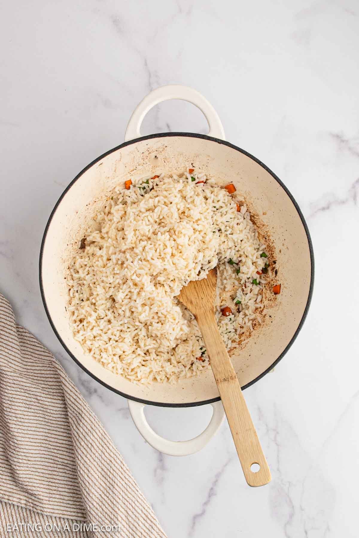 A white pot filled with delicious fried rice mixed with small pieces of vegetables, with a wooden spoon resting inside. The pot sits on a white marble surface next to a beige striped towel.