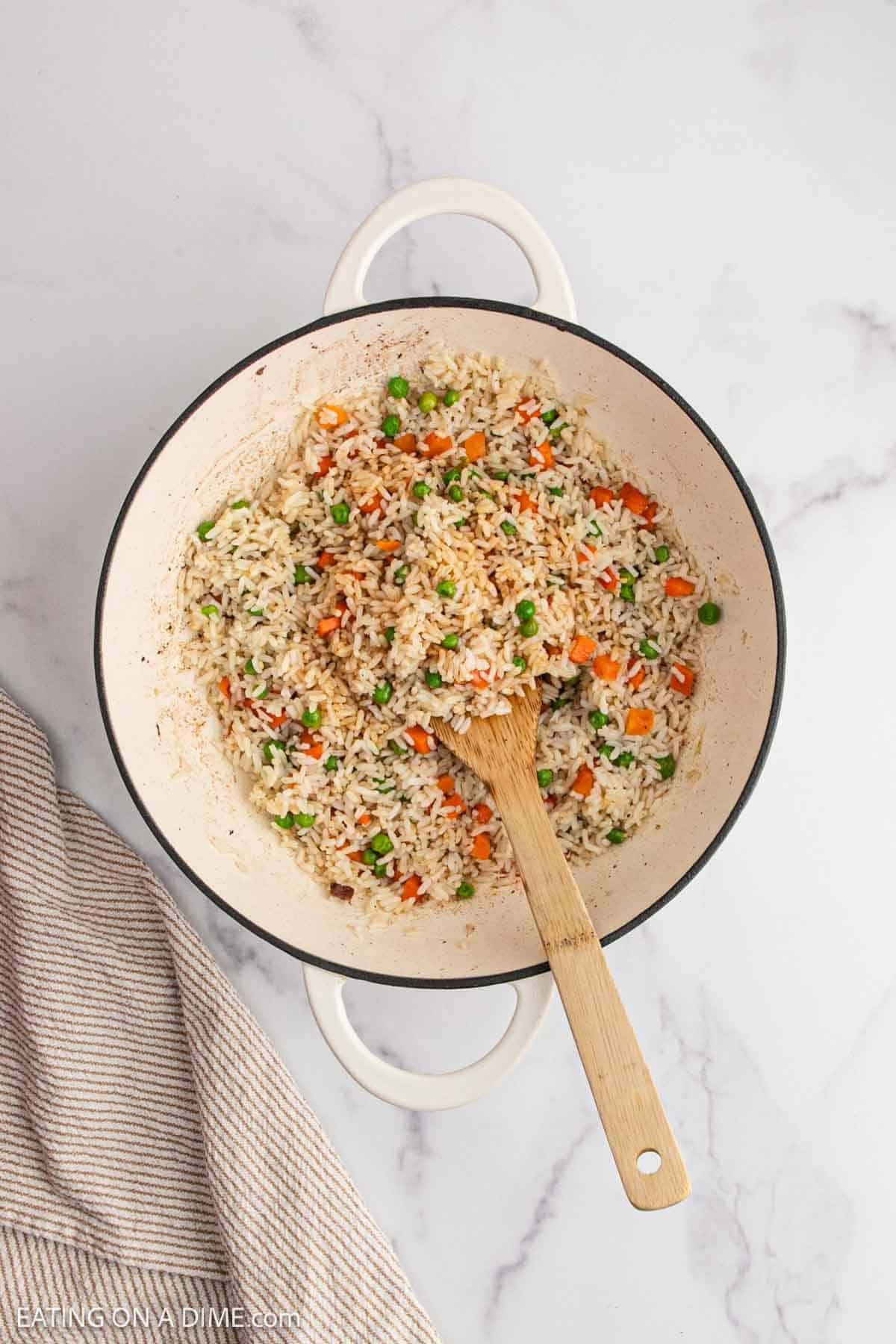 A white pot filled with cooked fried rice mixed with peas and diced carrots, with a wooden spoon resting inside. The pot is on a white marble surface next to a beige striped cloth.