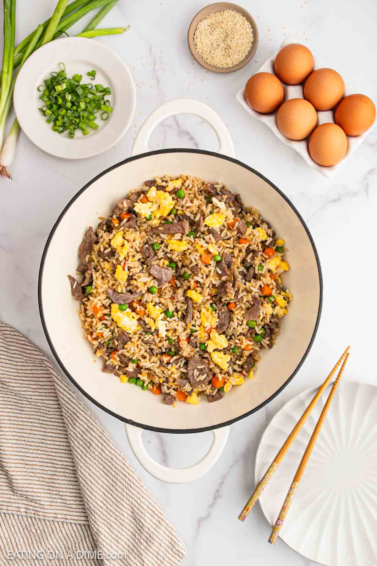 A white pot filled with Beef Fried Rice—scrambled eggs, beef, peas, and carrots—sits on a marble counter. Surrounding it are eggs, green onions, sesame seeds, a towel, an empty plate, and chopsticks.