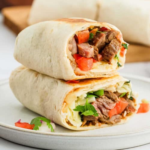 A close-up of carne asada burritos cut in half, stacked on a plate, filled with grilled meat, chopped tomatoes, fresh cilantro, and shredded lettuce wrapped in a flour tortilla.