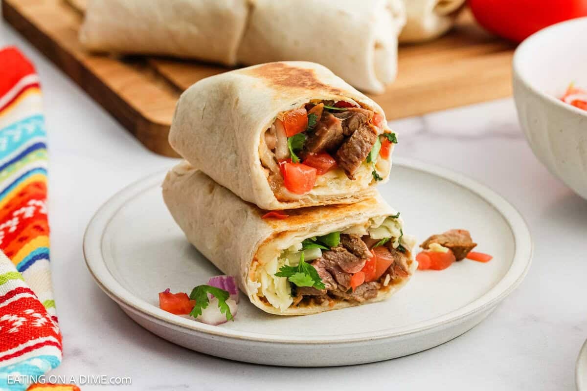 Two Carne Asada Burritos are stacked on a white plate, revealing their filling of grilled beef, diced tomatoes, lettuce, onions, and cilantro. A colorful striped cloth and more burritos are visible in the background.