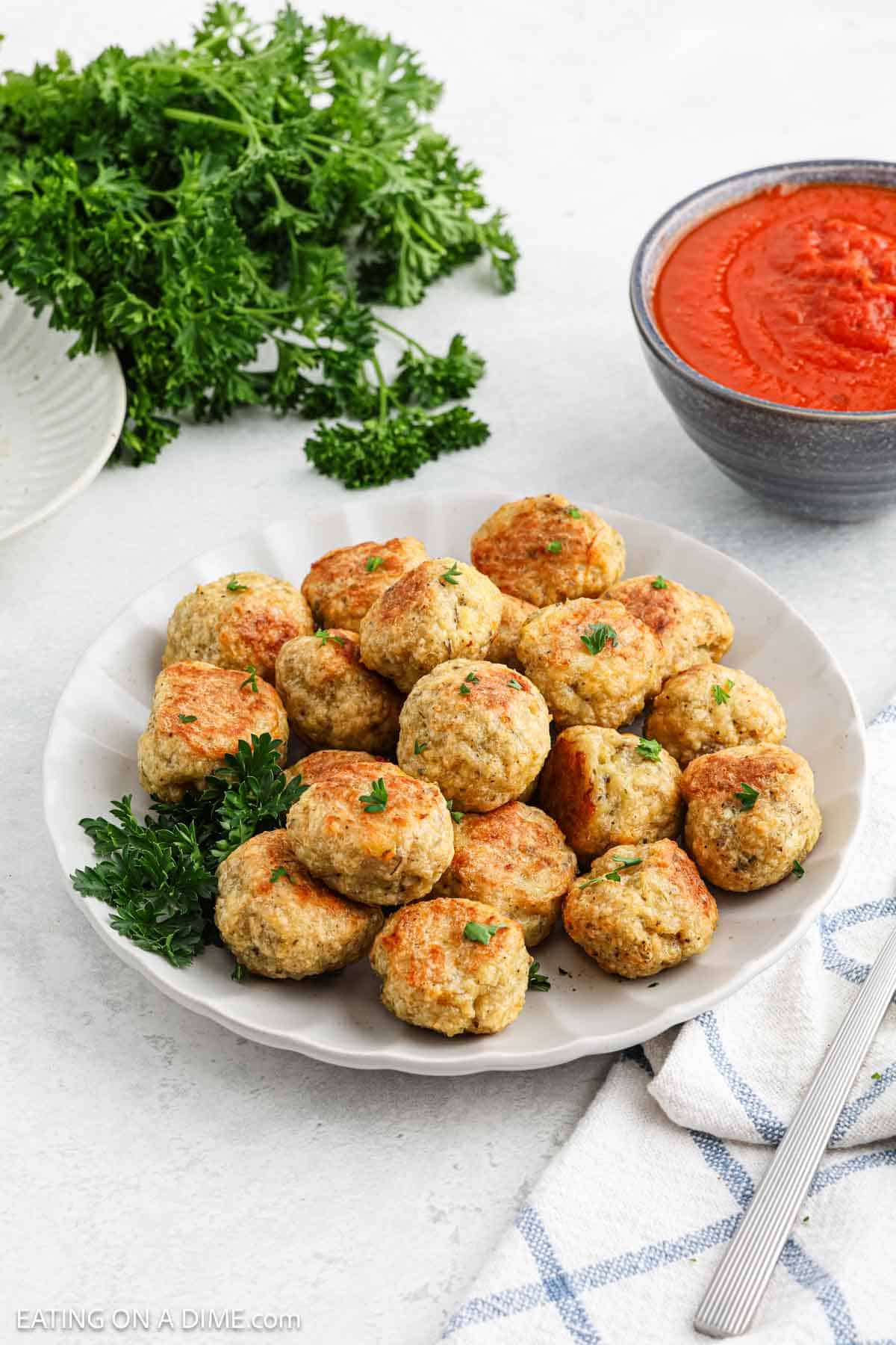 A plate of baked chicken meatballs garnished with parsley sits on a white surface next to a bowl of marinara sauce, fresh parsley, and a striped towel.
