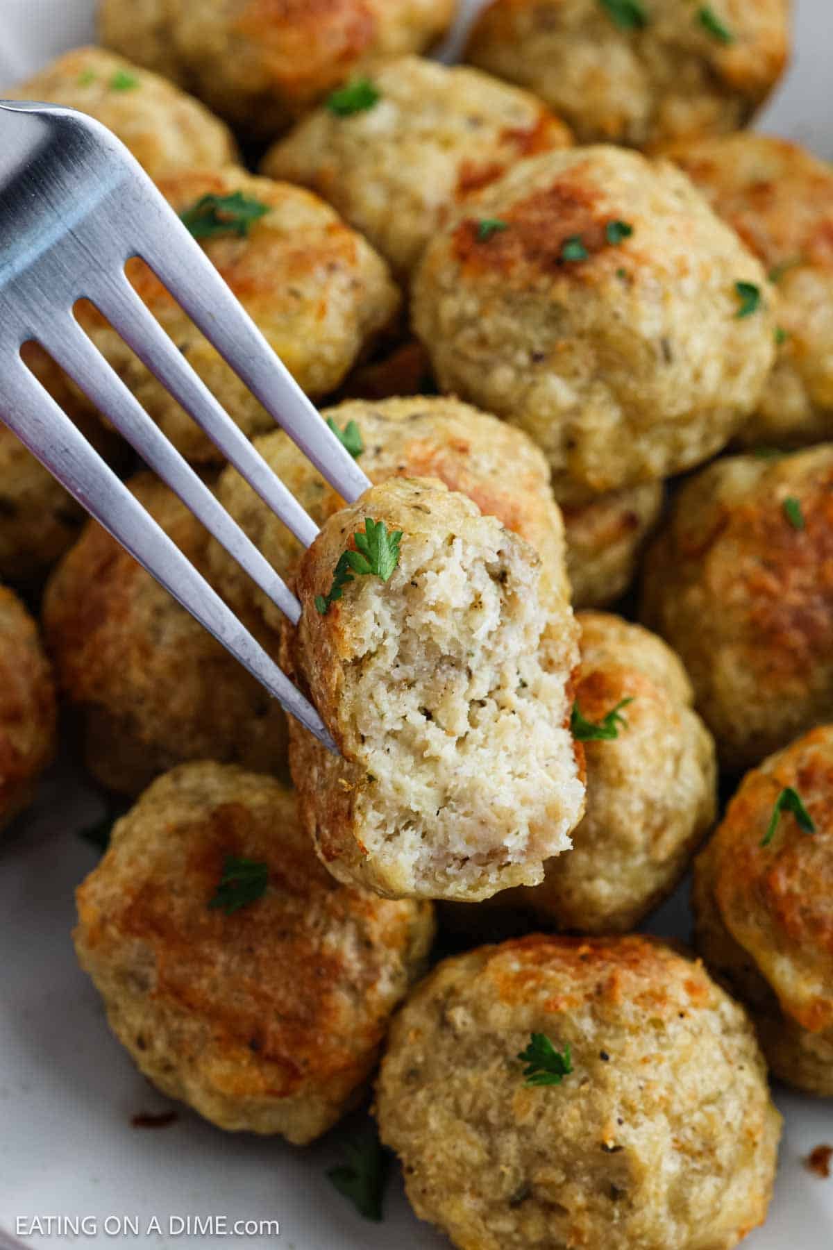 A fork holds a bitten Chicken Meatball above a plate full of more browned chicken meatballs, garnished with small pieces of parsley.