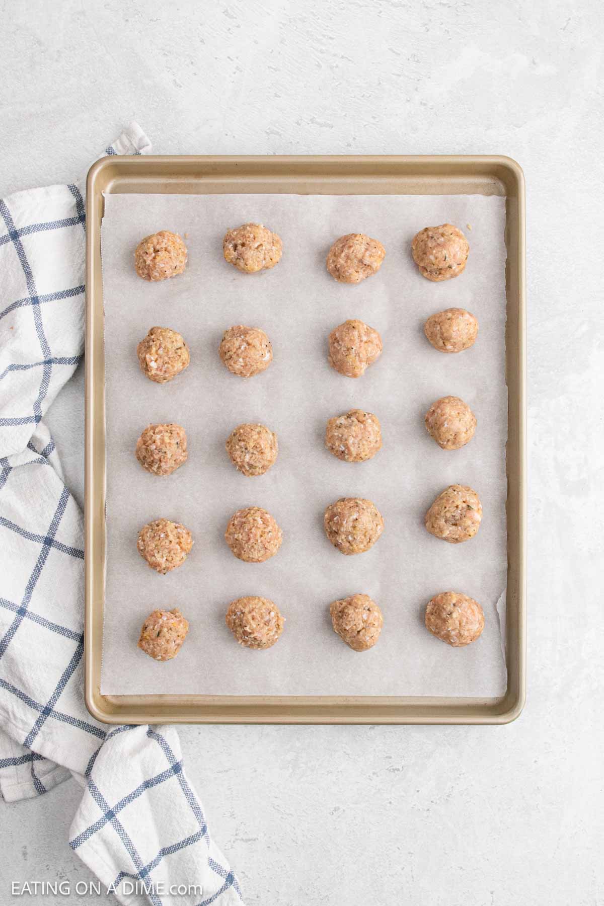 A baking sheet lined with parchment paper holds 20 evenly spaced, unbaked chicken meatballs. A blue and white checkered kitchen towel is placed to the side on a light countertop.