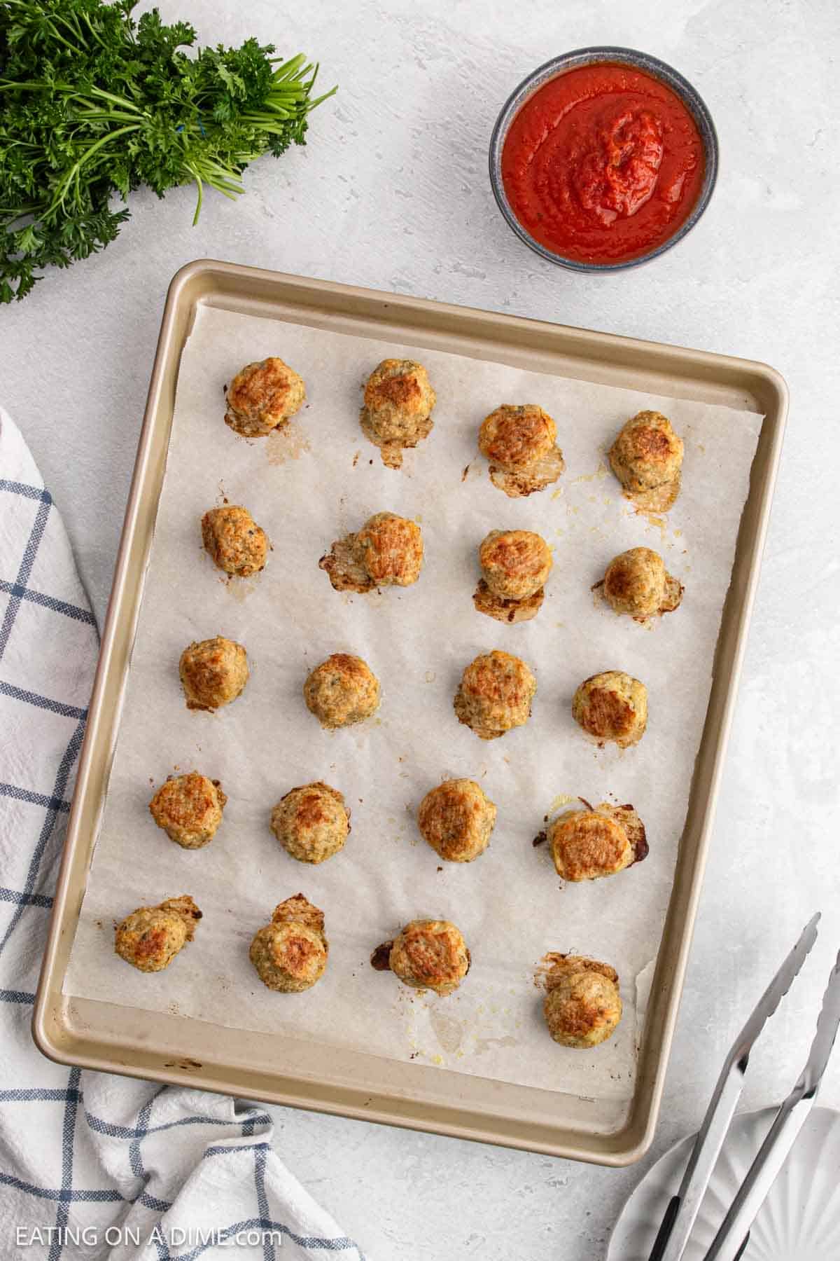 A baking sheet lined with parchment paper holds evenly spaced chicken meatballs. Beside it are a bowl of red tomato sauce, a bunch of fresh parsley, a kitchen towel, and metal tongs.