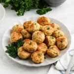 A white plate filled with golden-brown chicken meatballs garnished with fresh parsley, set on a light surface with a sprig of parsley and a striped cloth napkin nearby—perfect for anyone seeking an easy chicken meatball recipe.