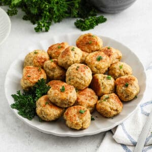 A white plate filled with golden-brown chicken meatballs garnished with fresh parsley, set on a light surface with a sprig of parsley and a striped cloth napkin nearby—perfect for anyone seeking an easy chicken meatball recipe.