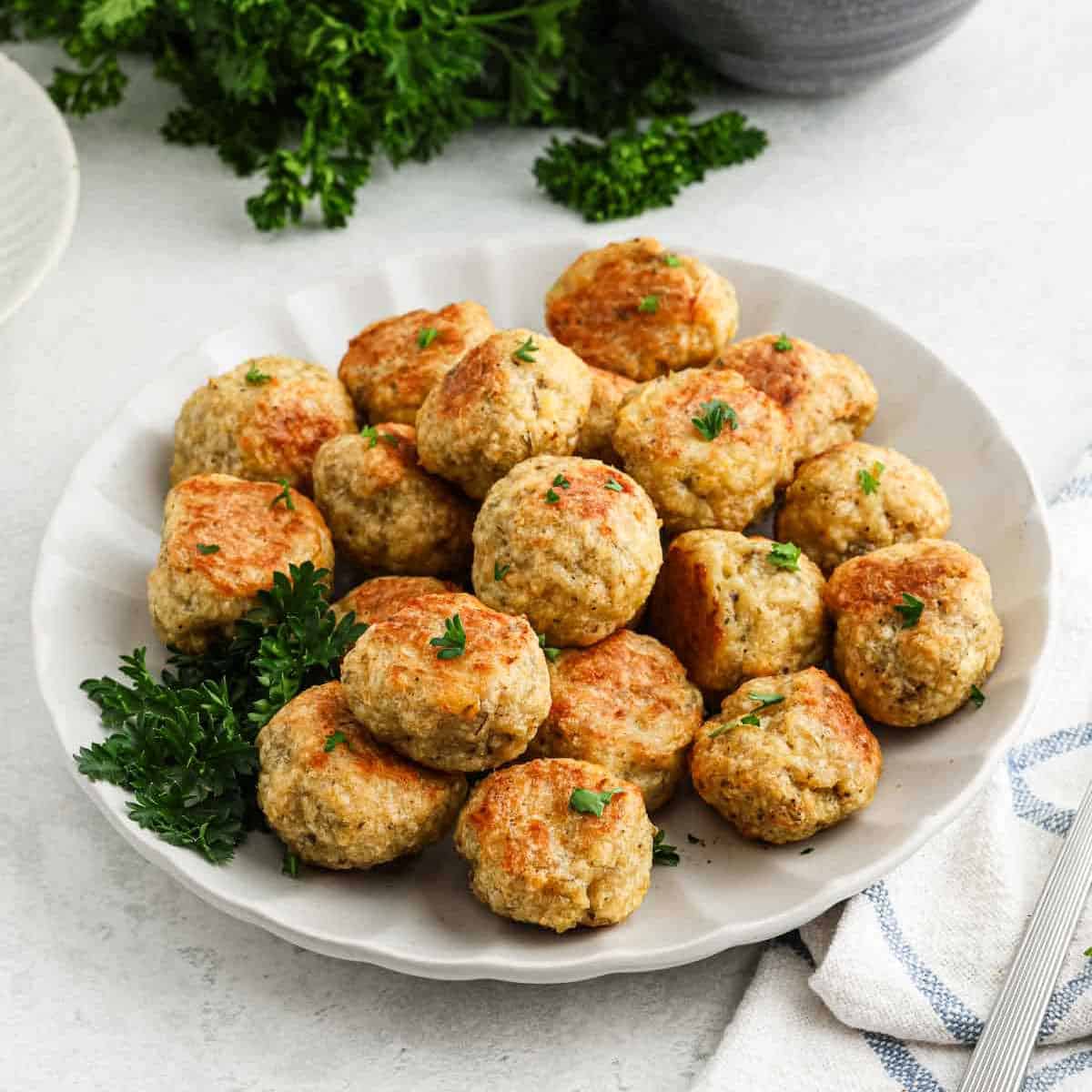 A white plate filled with golden-brown chicken meatballs garnished with fresh parsley, set on a light surface with a sprig of parsley and a striped cloth napkin nearby—perfect for anyone seeking an easy chicken meatball recipe.