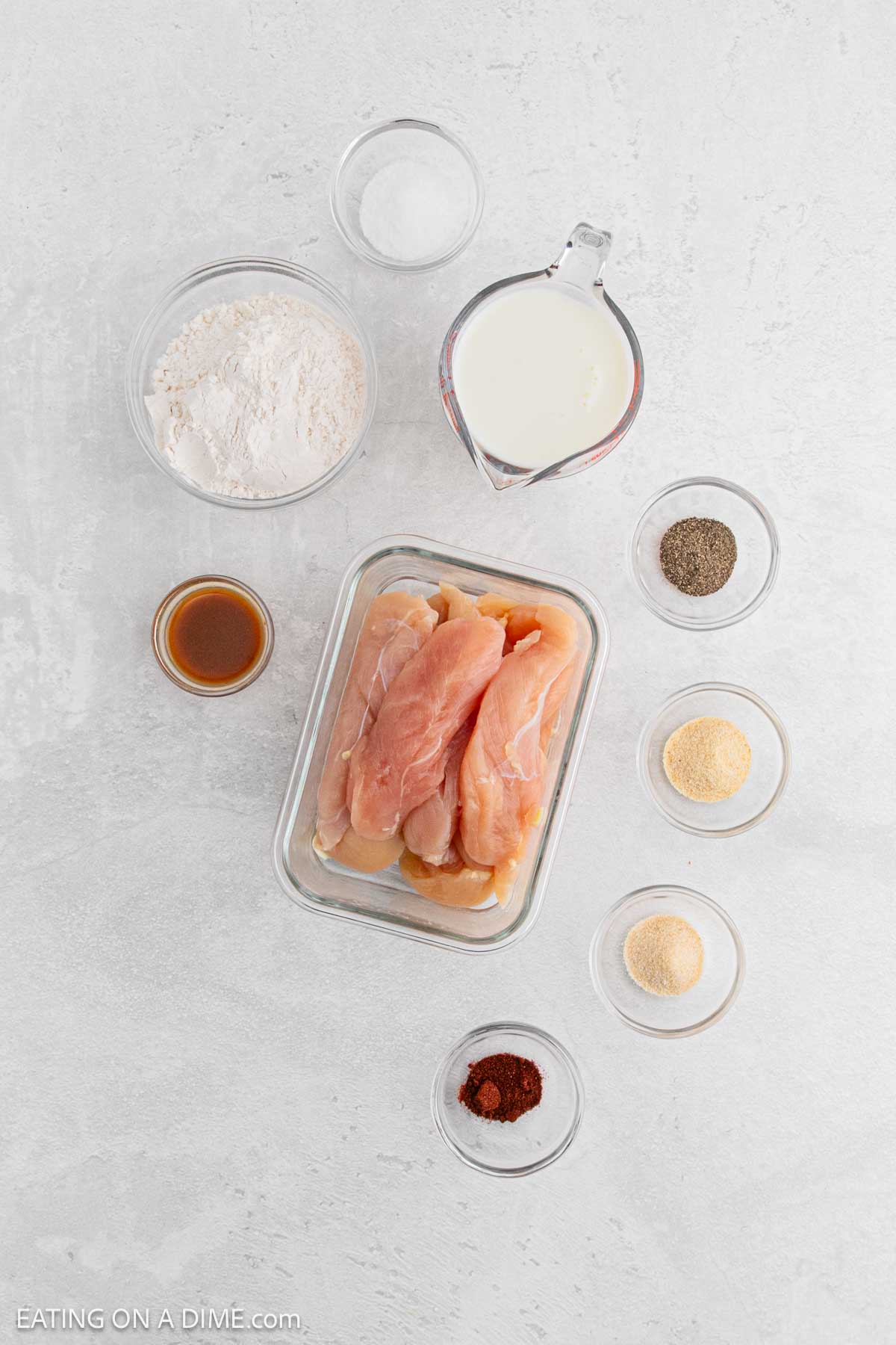 Raw chicken breasts in a glass container, surrounded by small bowls of flour, milk, seasonings, and a small bowl of brown liquid—perfect ingredients for making classic chicken and waffles on a light countertop.