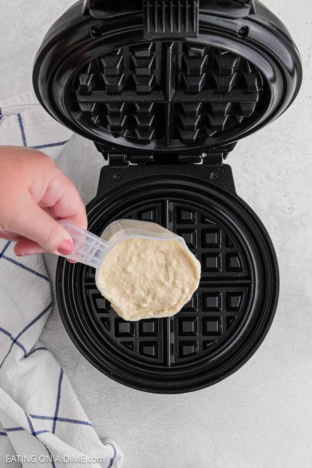 A hand pours waffle batter from a measuring cup onto the center of an open, preheated waffle maker—perfect for making classic chicken and waffles. A white and blue striped dish towel is nearby on the counter.