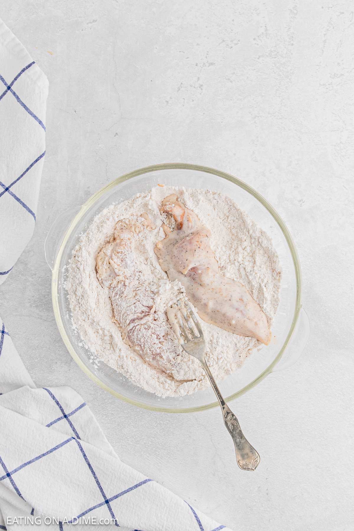 Two raw chicken breasts being coated in flour in a glass bowl with a fork, on a light-colored countertop next to a white and blue checkered kitchen towel—perfect for making classic Chicken and Waffles.