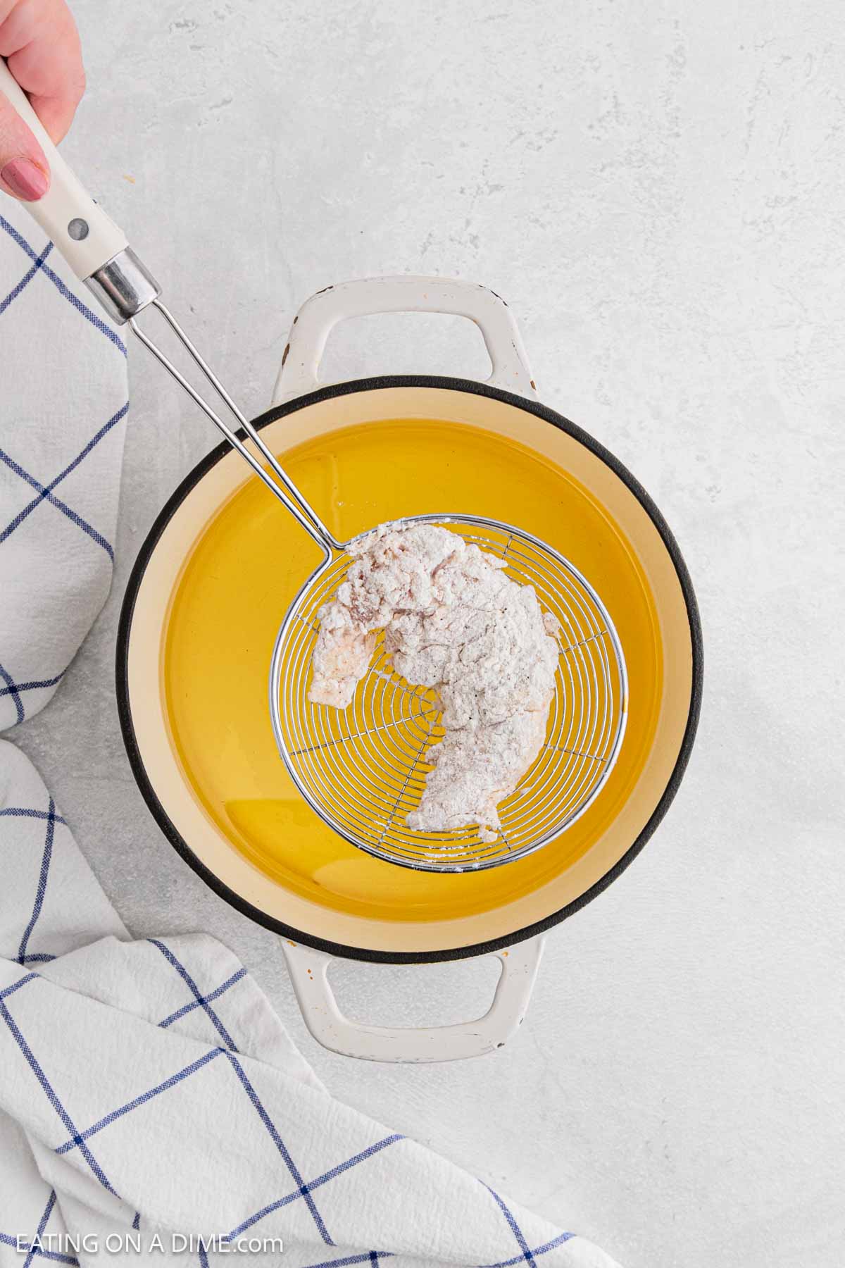 A hand holds a wire skimmer with a breaded chicken strip above a yellow pot of oil, ready to fry for delicious Chicken and Waffles. A white and blue kitchen towel rests nearby on a light surface.