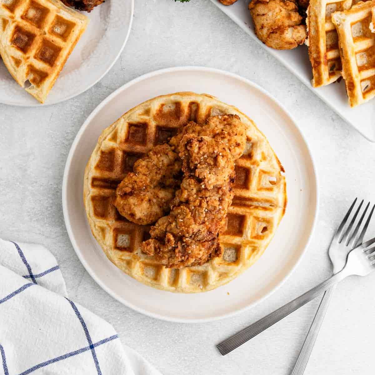 A plate with a large waffle topped with two pieces of crispy fried chicken—a classic Chicken and Waffles dish. Nearby are forks, a blue-and-white napkin, and extra plates featuring more chicken recipes. The background is a light, textured surface.