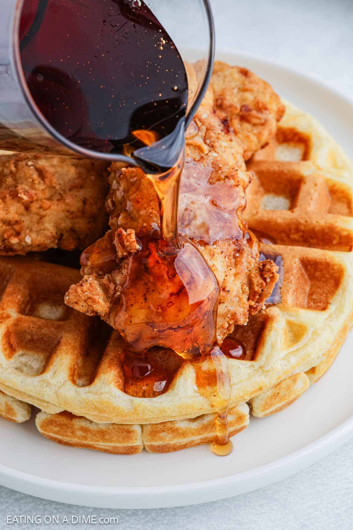 A close-up of syrup being poured over crispy Chicken and Waffles, with golden fried chicken pieces stacked on top of fluffy waffles on a white plate.