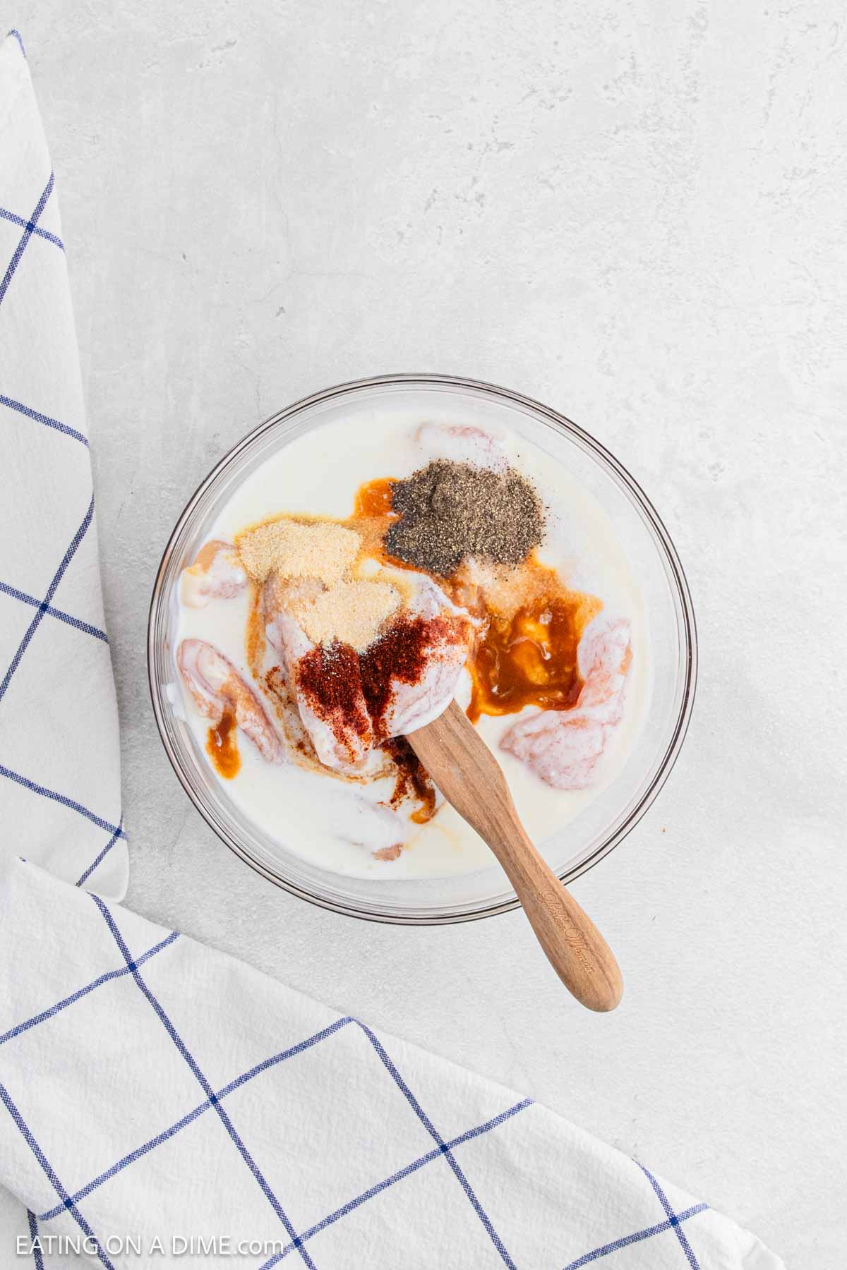 A glass bowl filled with raw chicken pieces, creamy liquid, and various spices sits on a light surface beside a white cloth with blue grid lines—perfect for prepping classic Chicken and Waffles. A wooden spoon rests in the mixture.