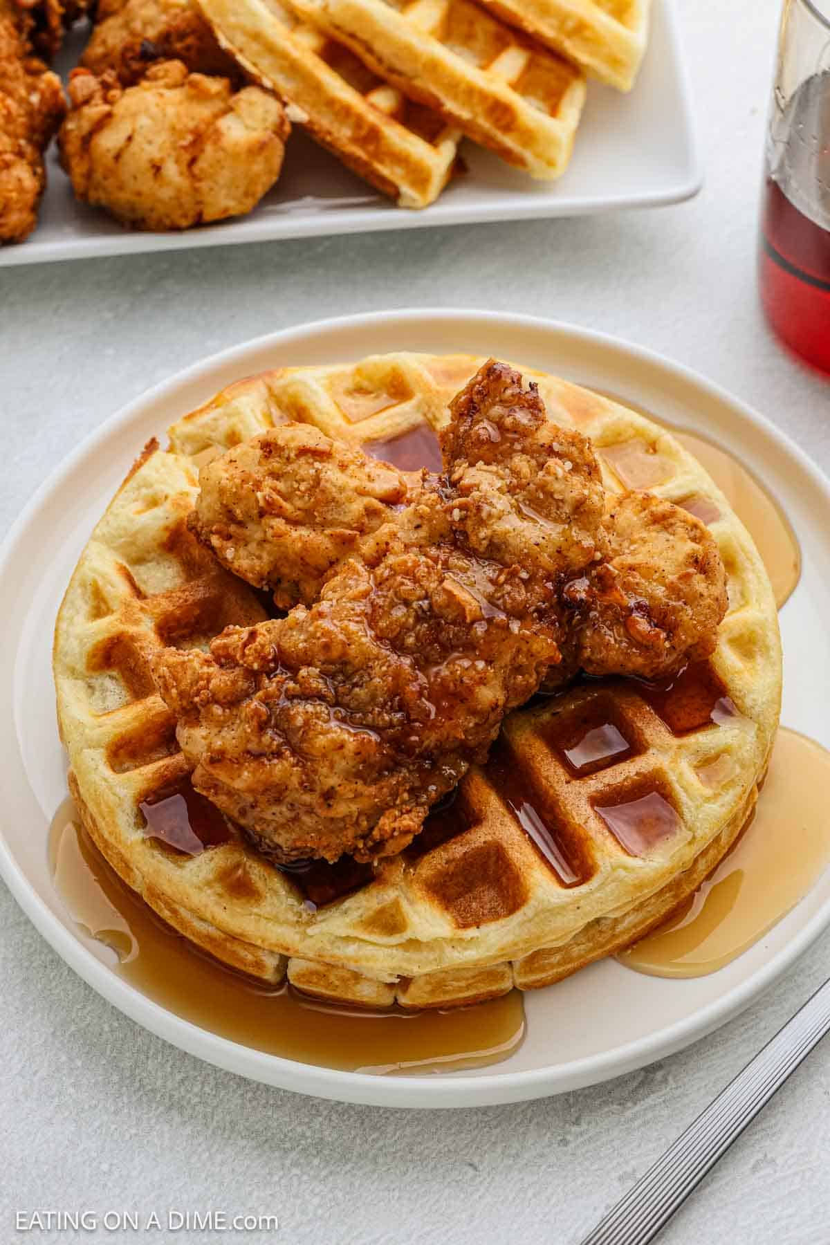 A plate with a golden waffle topped with crispy fried chicken and drizzled with syrup—classic chicken and waffles. A fork rests beside the plate, with more waffles, chicken, and syrup in the background.