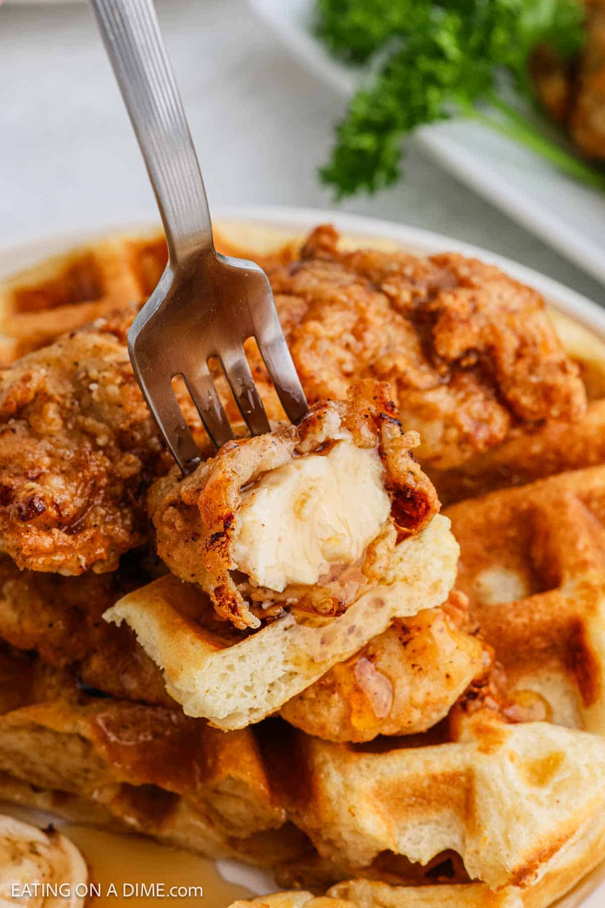 A close-up of a fork holding a bite of crispy chicken and waffles, with more golden chicken and waffles stacked underneath on a plate. Fresh parsley is blurred in the background.