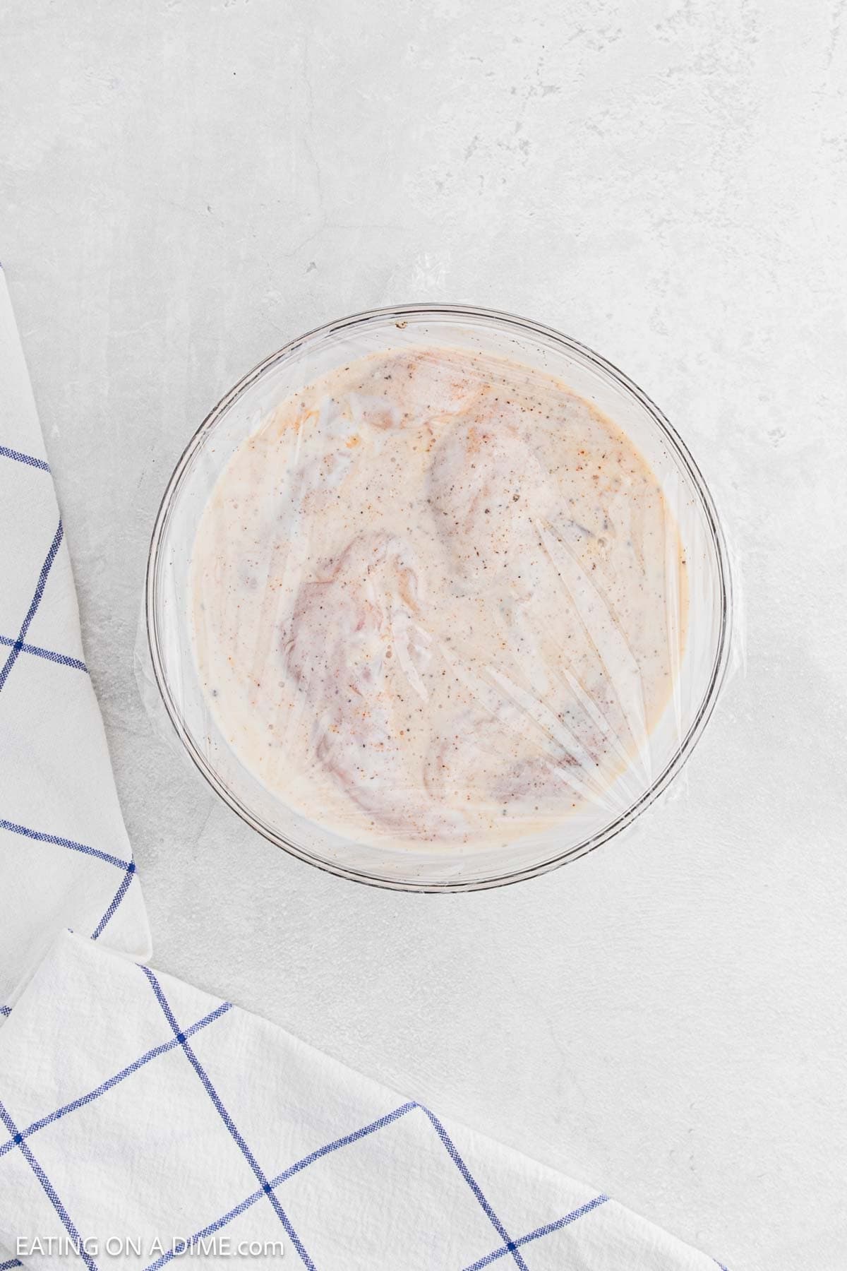 A glass bowl filled with raw chicken marinating in a creamy, seasoned mixture—perfect for Chicken and Waffles—covered with plastic wrap. The bowl rests on a light countertop next to a white towel with blue stripes.