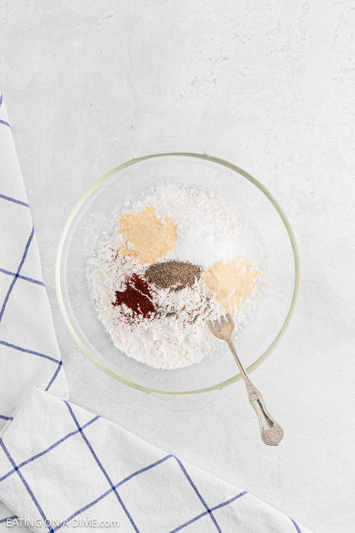 A glass bowl filled with flour and various spices for chicken and waffles, including black pepper, paprika, and garlic powder, sits on a white surface next to a white towel with blue grid lines. A fork rests inside the bowl.
