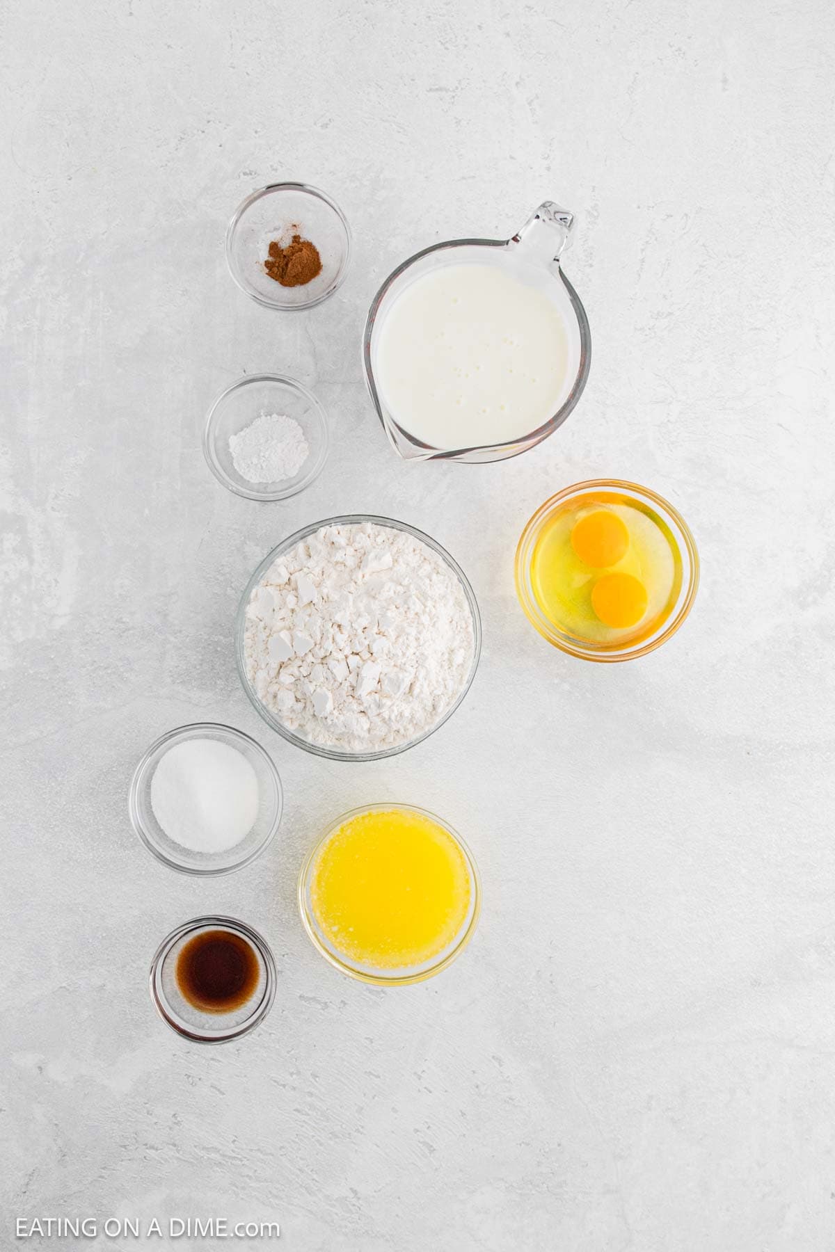 Top-down view of measured baking ingredients in clear bowls—flour, sugar, melted butter, vanilla extract, milk, two eggs, cinnamon, and baking powder—neatly arranged on a light surface for homemade waffles or chicken and waffles.