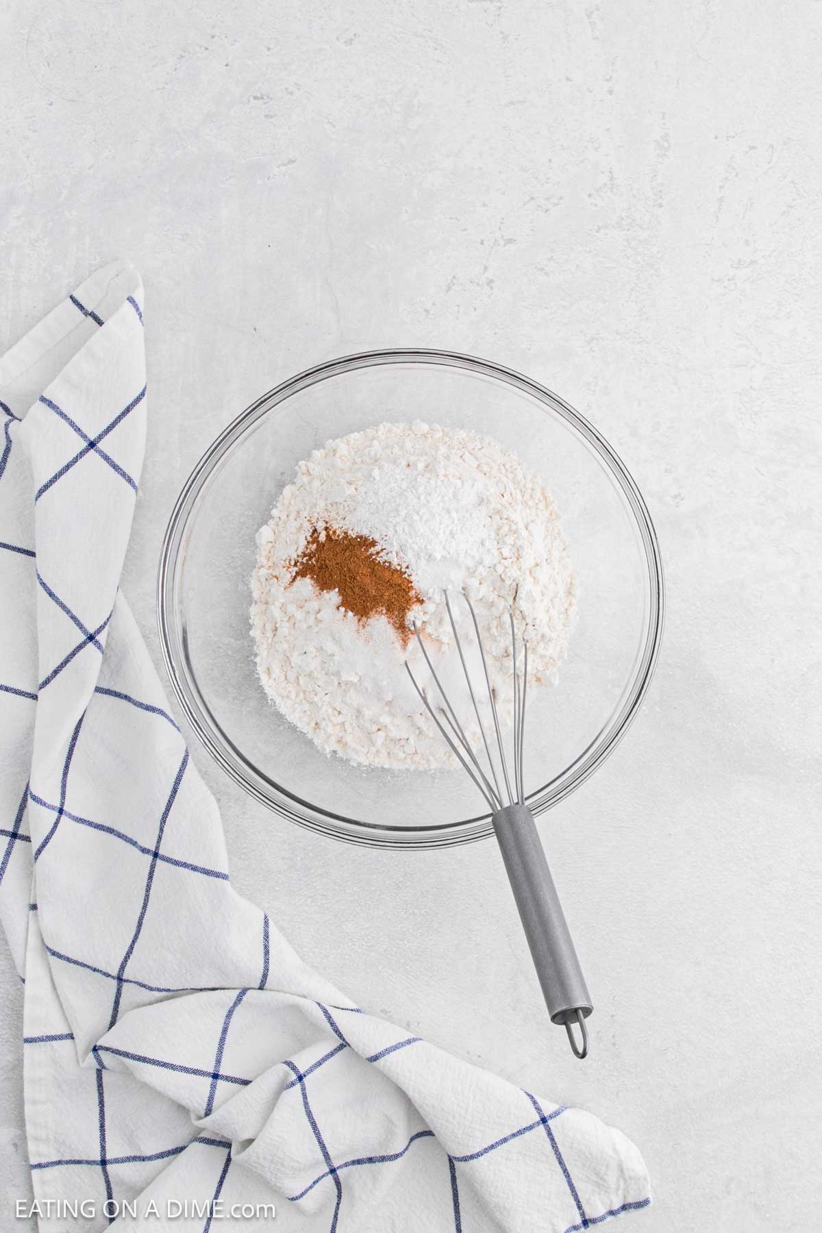 A glass mixing bowl filled with flour, sugar, and cinnamon—ingredients perfect for chicken and waffles—sits on a white surface with a metal whisk inside. A white kitchen towel with blue grid lines is draped beside the bowl.