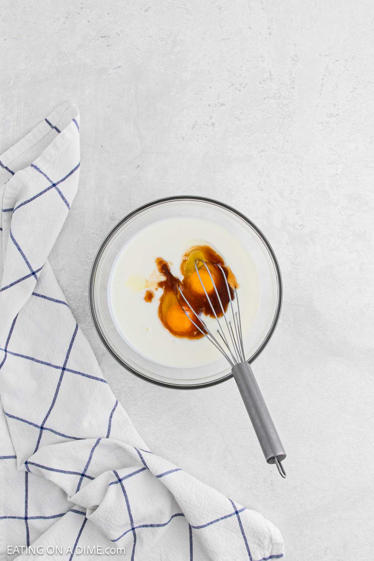 A glass bowl with white liquid, likely milk, and brown liquid, possibly vanilla extract, being whisked together—perfect for a breakfast recipe like Chicken and Waffles. The bowl sits on a light countertop beside a white kitchen towel with blue lines.