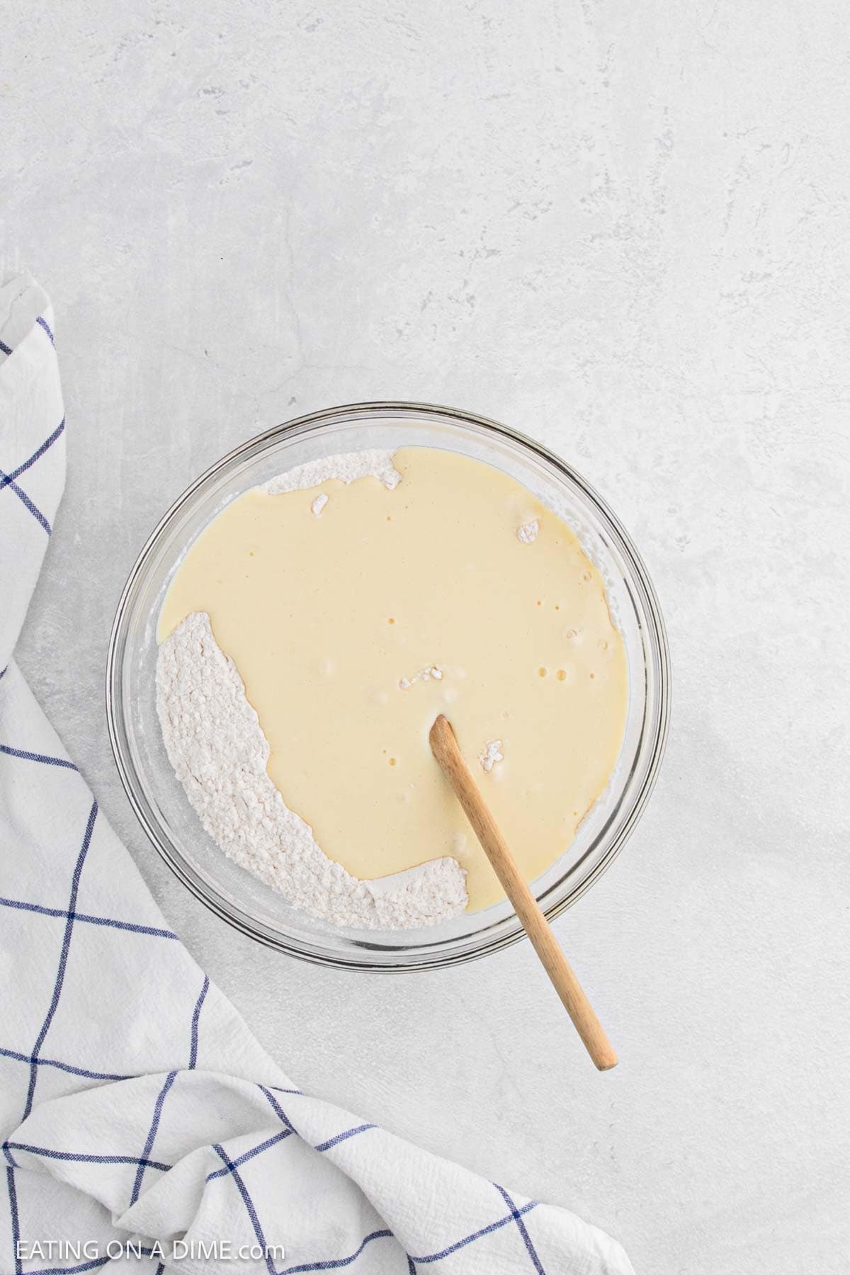 A glass bowl with flour and batter for chicken and waffles is being mixed with a wooden spoon on a light gray surface, next to a white kitchen towel with blue stripes.
