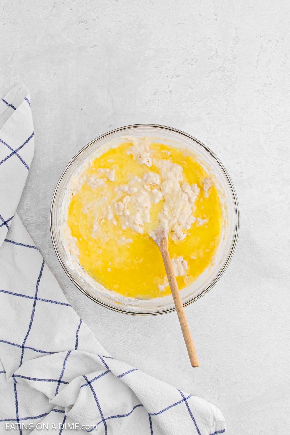 A glass bowl with a mixture of wet and dry ingredients for chicken and waffles being stirred with a wooden spoon, next to a white towel with blue grid lines on a light surface.