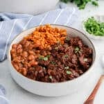 A white bowl filled with Mexican rice, refried beans, and beef Chile Colorado chili garnished with chopped cilantro, placed on a light surface next to a blue-striped napkin and a small dish of herbs.