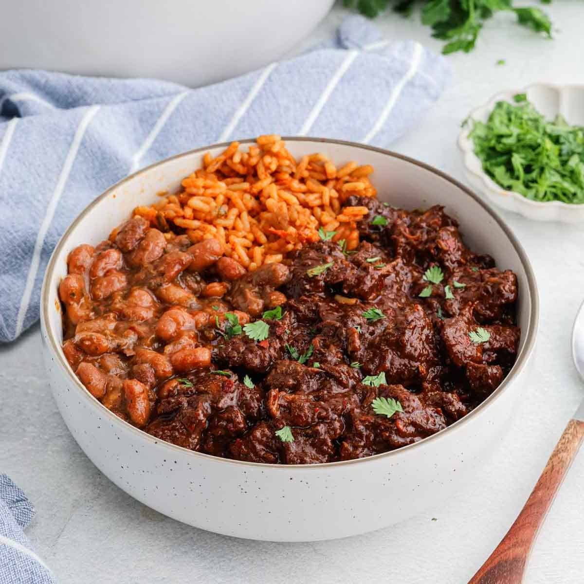 A white bowl filled with Mexican rice, refried beans, and beef Chile Colorado chili garnished with chopped cilantro, placed on a light surface next to a blue-striped napkin and a small dish of herbs.