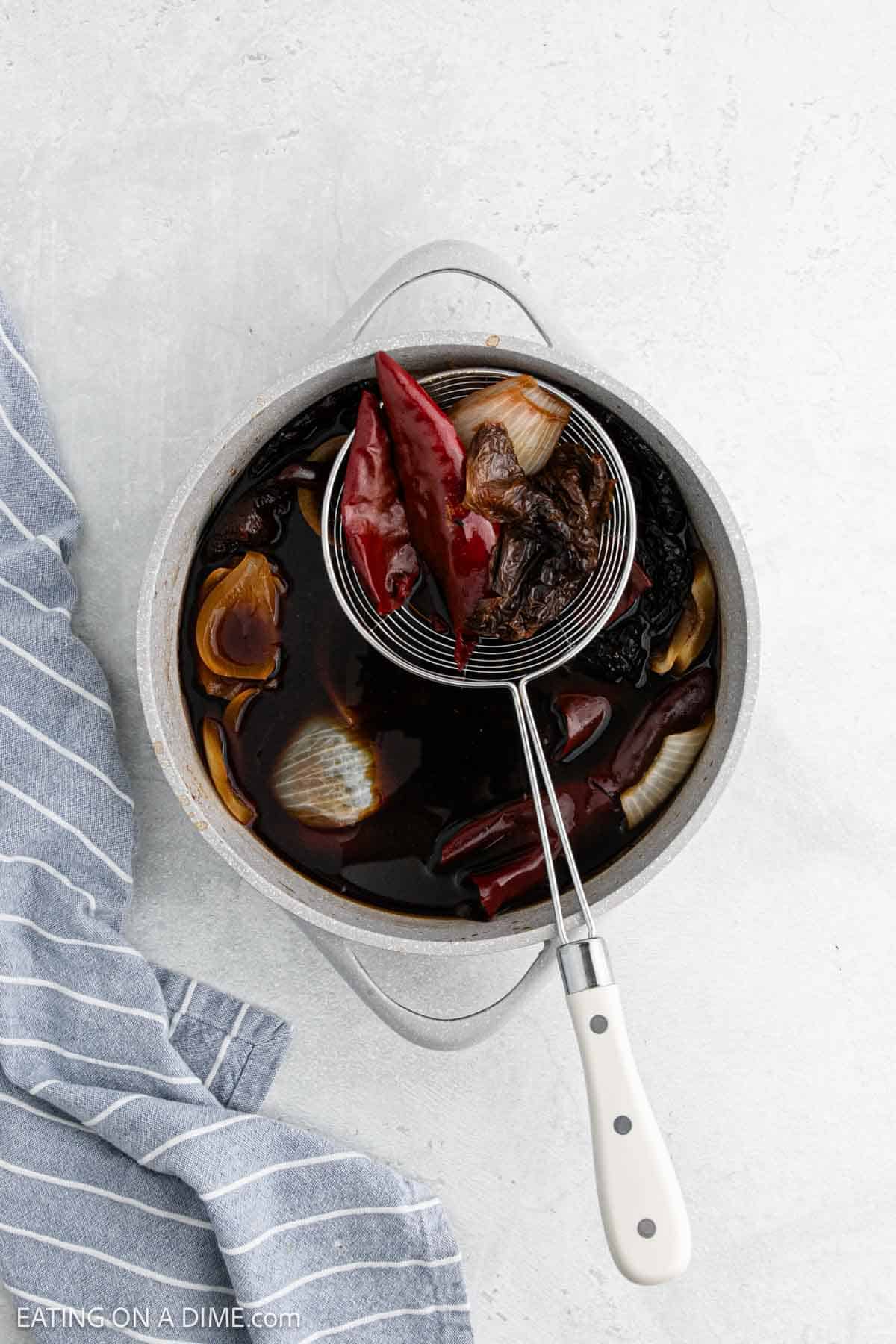 A pot of rich Chile Colorado broth with halved onions, dried chili peppers, and vegetables simmers. A slotted spoon with a white handle lifts the ingredients above the pot. A blue striped towel lies nearby.