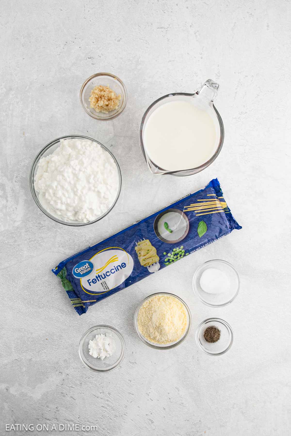 Ingredients for homemade Cottage Cheese Alfredo sauce are arranged on a light surface, including cottage cheese, fettuccine pasta, milk, grated Parmesan, garlic, salt, pepper, and cornstarch in various bowls and containers.