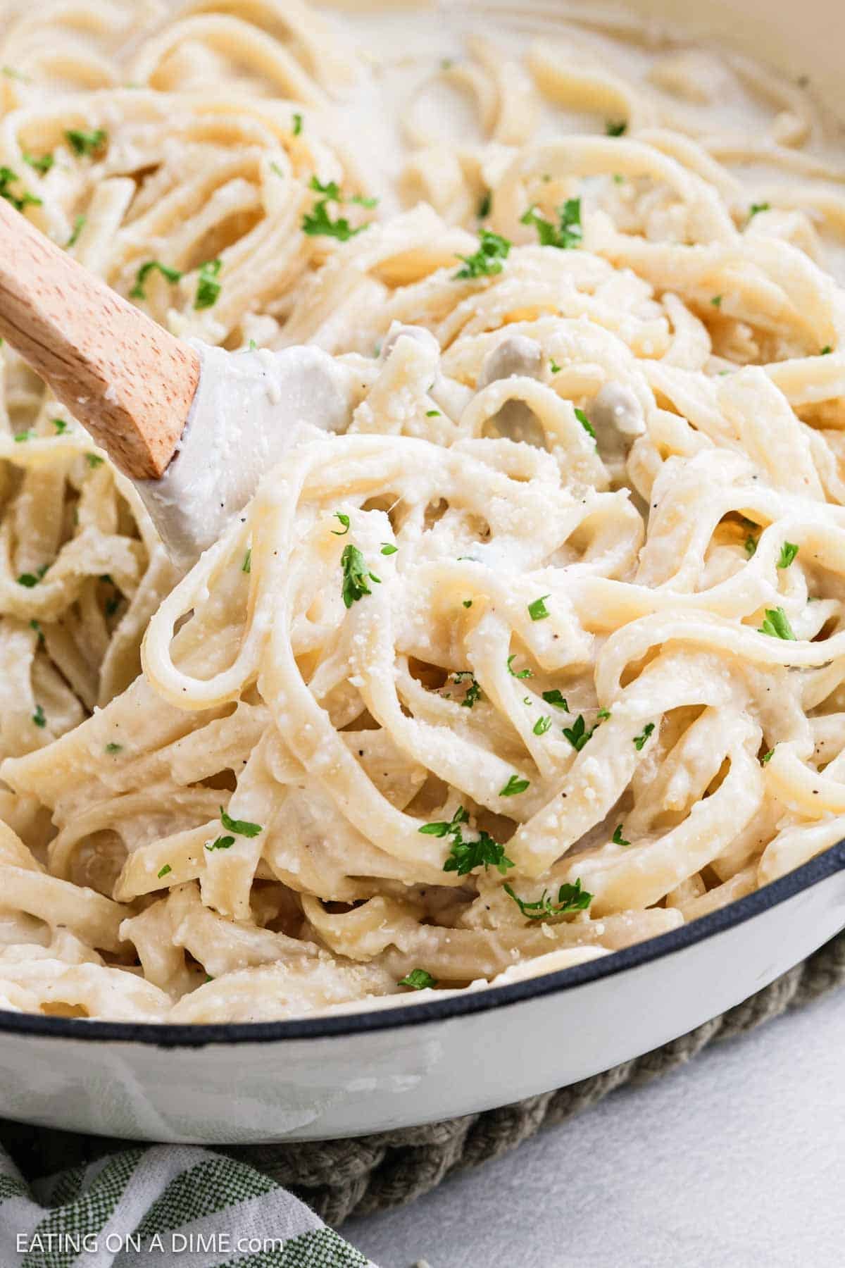 A close-up of creamy fettuccine coated in rich alfredo sauce being stirred in a white pan, topped with chopped parsley and grated Parmesan cheese.