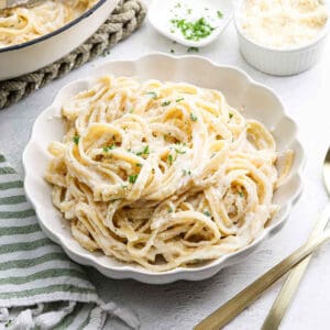 A white bowl filled with creamy Cottage Cheese Alfredo, garnished with chopped parsley and grated Parmesan cheese, sits on a white table next to a gold fork and knife, with a striped napkin nearby.