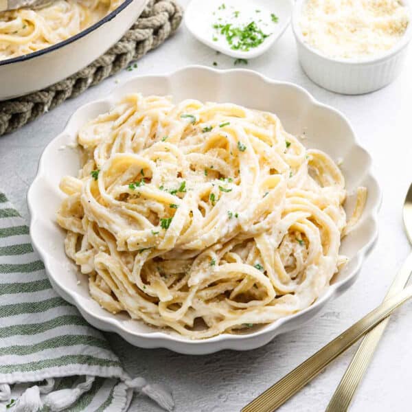 A white bowl filled with creamy Cottage Cheese Alfredo, garnished with chopped parsley and grated Parmesan cheese, sits on a white table next to a gold fork and knife, with a striped napkin nearby.