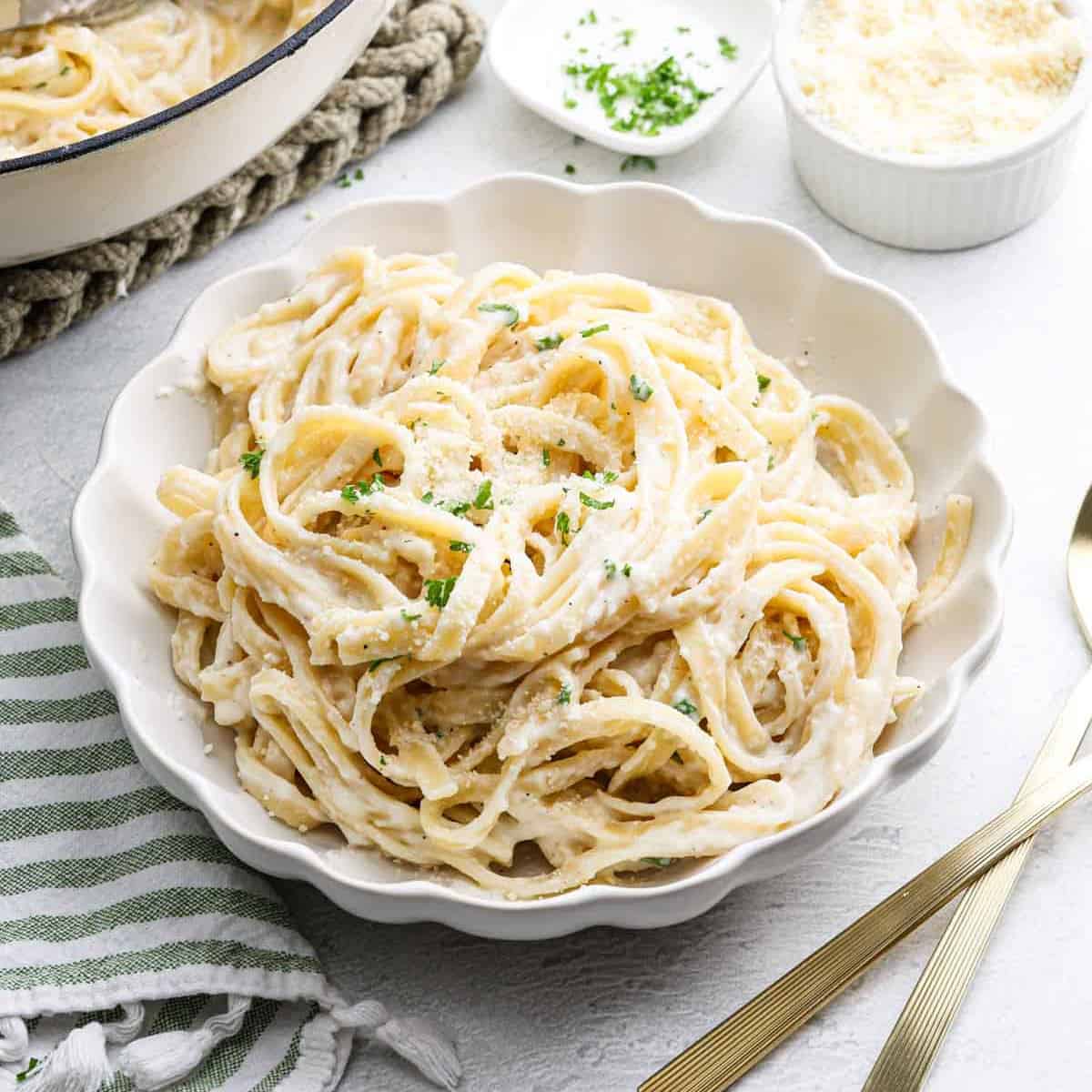 A white bowl filled with creamy Cottage Cheese Alfredo, garnished with chopped parsley and grated Parmesan cheese, sits on a white table next to a gold fork and knife, with a striped napkin nearby.
