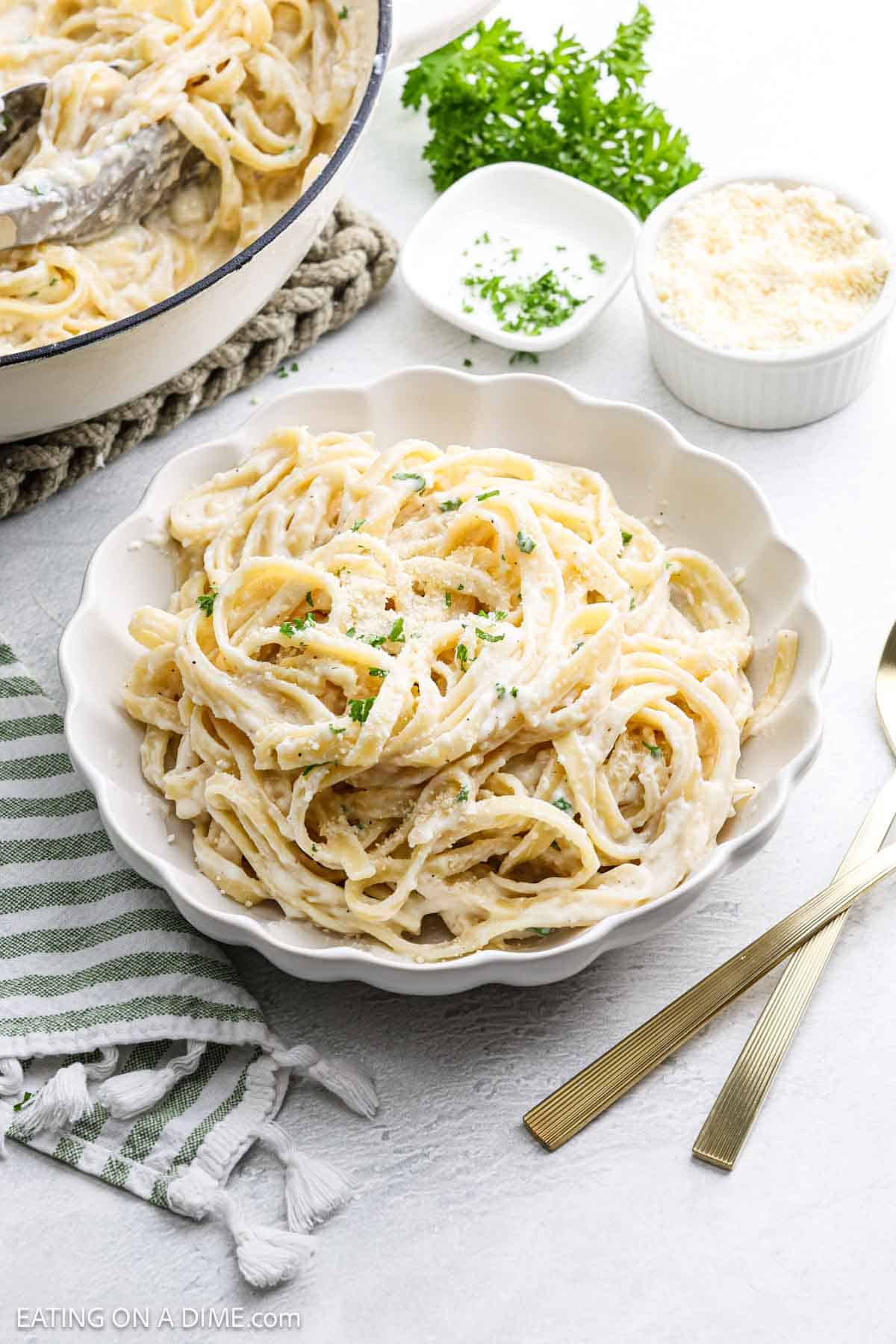 A bowl of creamy Cottage Cheese Alfredo garnished with chopped parsley sits on a white table next to gold utensils, a green striped napkin, and small bowls of grated cheese and herbs.