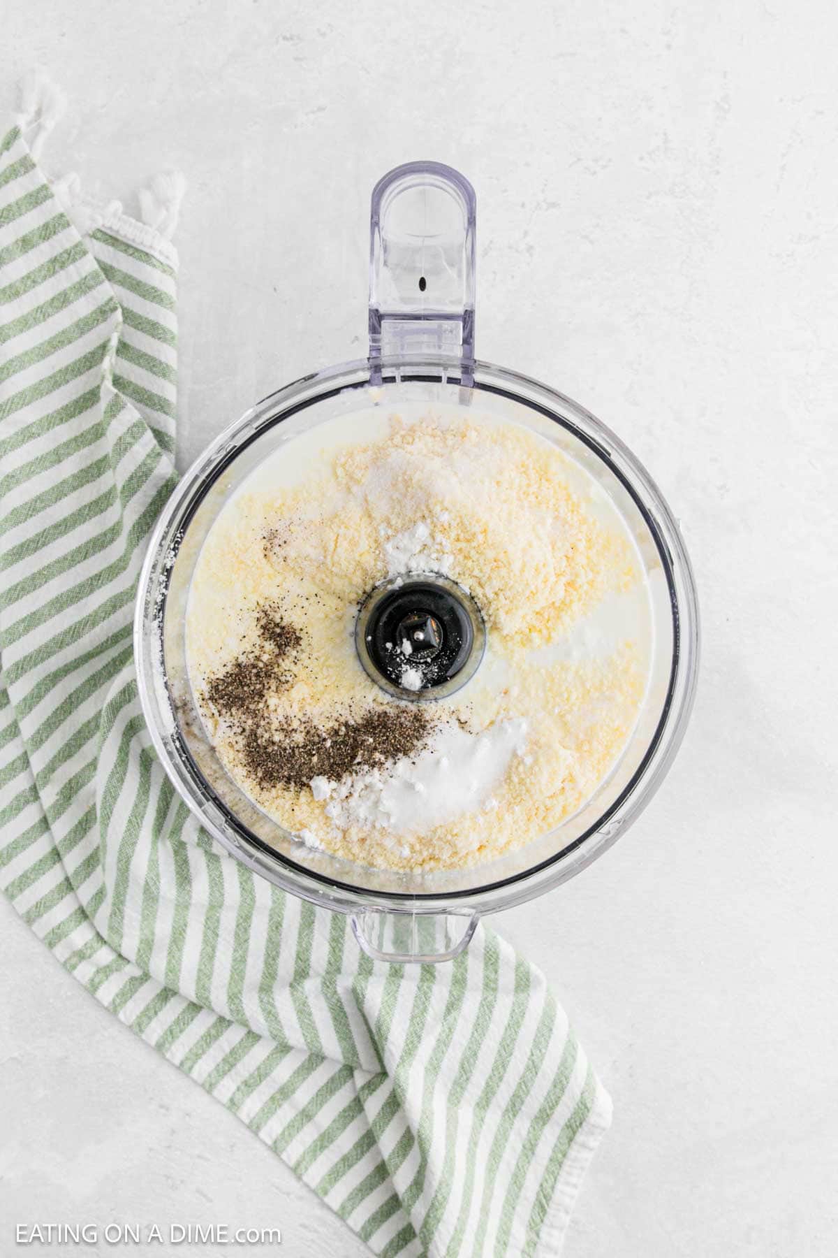 A food processor filled with cornmeal, milk, cottage cheese, and spices sits on a light surface next to a green and white striped kitchen towel.