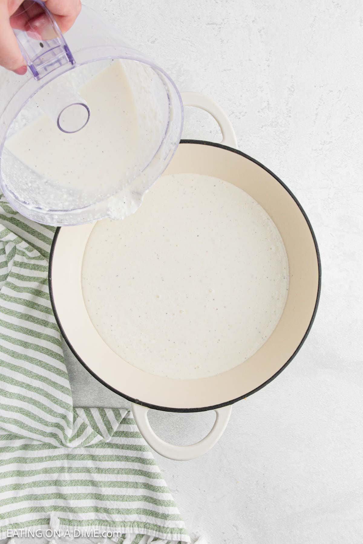 A creamy Cottage Cheese Alfredo sauce is being poured from a blender into a large white pot on a light surface, with a green and white striped towel nearby.