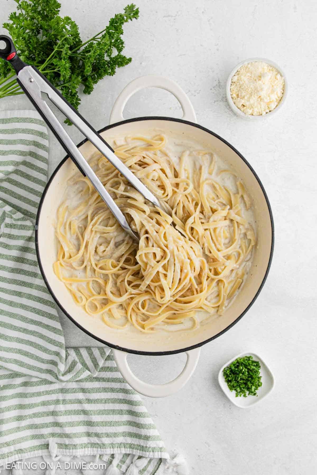 A pot of creamy Cottage Cheese Alfredo topped with tongs sits on a light surface, surrounded by a bowl of grated cheese, chopped parsley, and a striped green kitchen towel. Fresh parsley is in the upper left corner.
