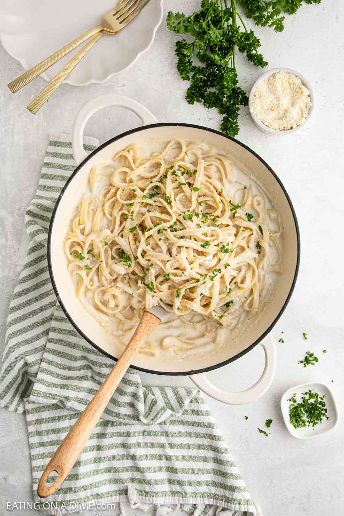 A white pot of creamy Cottage Cheese Alfredo garnished with chopped parsley, with a wooden spoon resting inside. Nearby are a striped towel, a dish of grated cheese, fresh parsley, and two gold forks on a white plate.