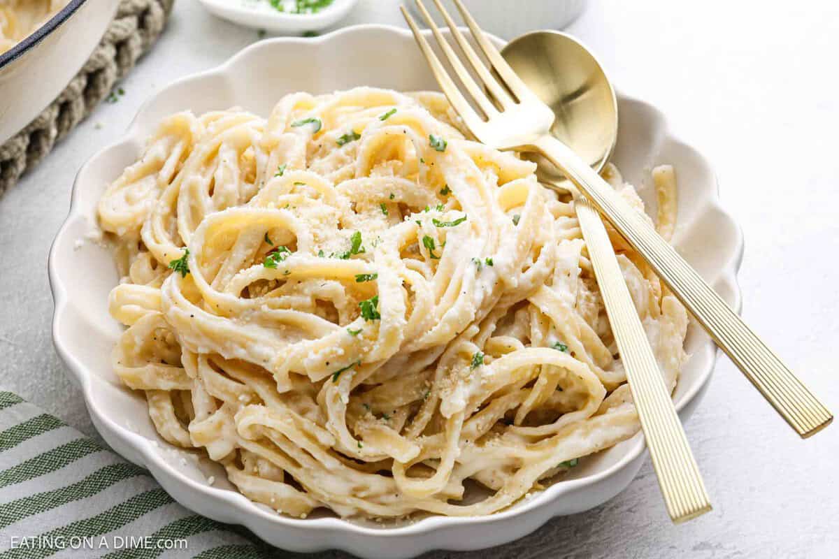 A white bowl filled with creamy Cottage Cheese Alfredo pasta, garnished with grated parmesan cheese and chopped parsley. A gold fork and spoon rest on the side of the bowl.