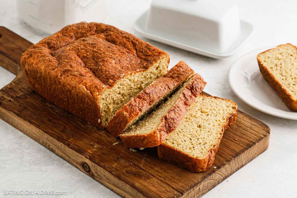 A loaf of sliced homemade Cottage Cheese Bread sits on a wooden cutting board, with three slices partially fanned out. A butter dish and an extra slice of bread on a plate are visible in the background.