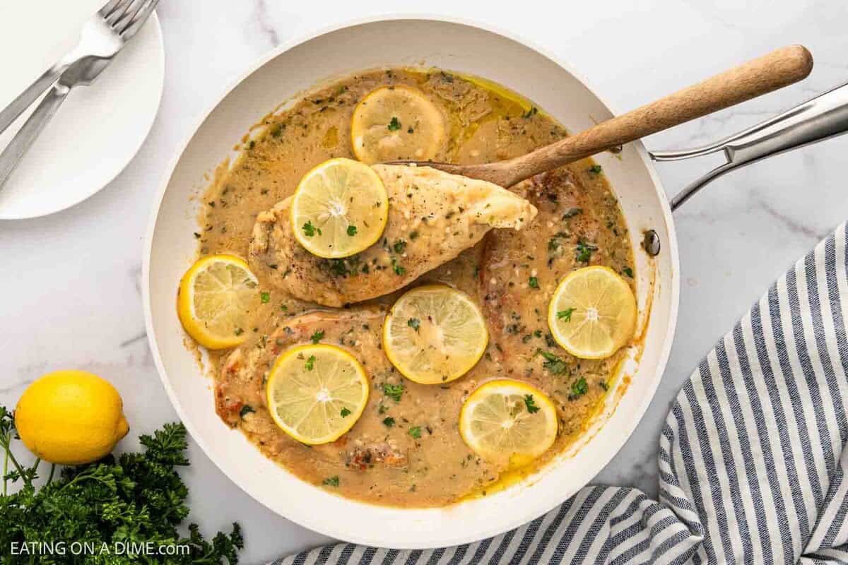 Chicken breast placed in a skillet covered with a creamy sauce and lemon slices with a serving on a wooden spoon