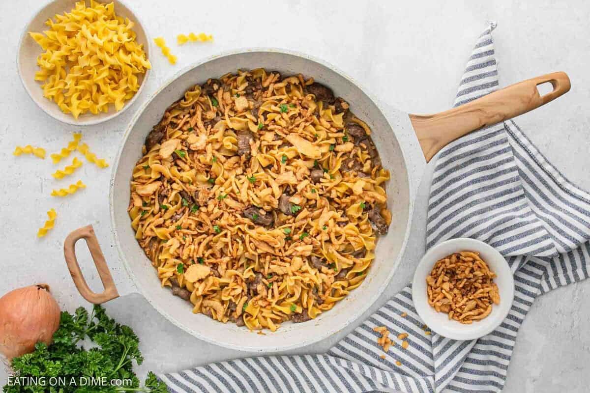 A skillet of French Onion Beef and noodle casserole topped with crispy fried onions sits on a striped cloth. Nearby are a bowl of uncooked noodles, a bowl of crispy onions, fresh parsley, and a whole onion. One pot dinner