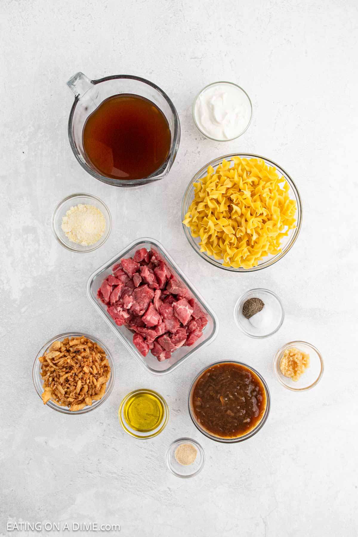 Flat lay of ingredients for Beef and Noodles on a white surface, featuring uncooked egg noodles, cubed beef, French Onion flavors with fried onions, beef broth, sour cream, gravy, spices, grated cheese, minced garlic, olive oil, and ground pepper.