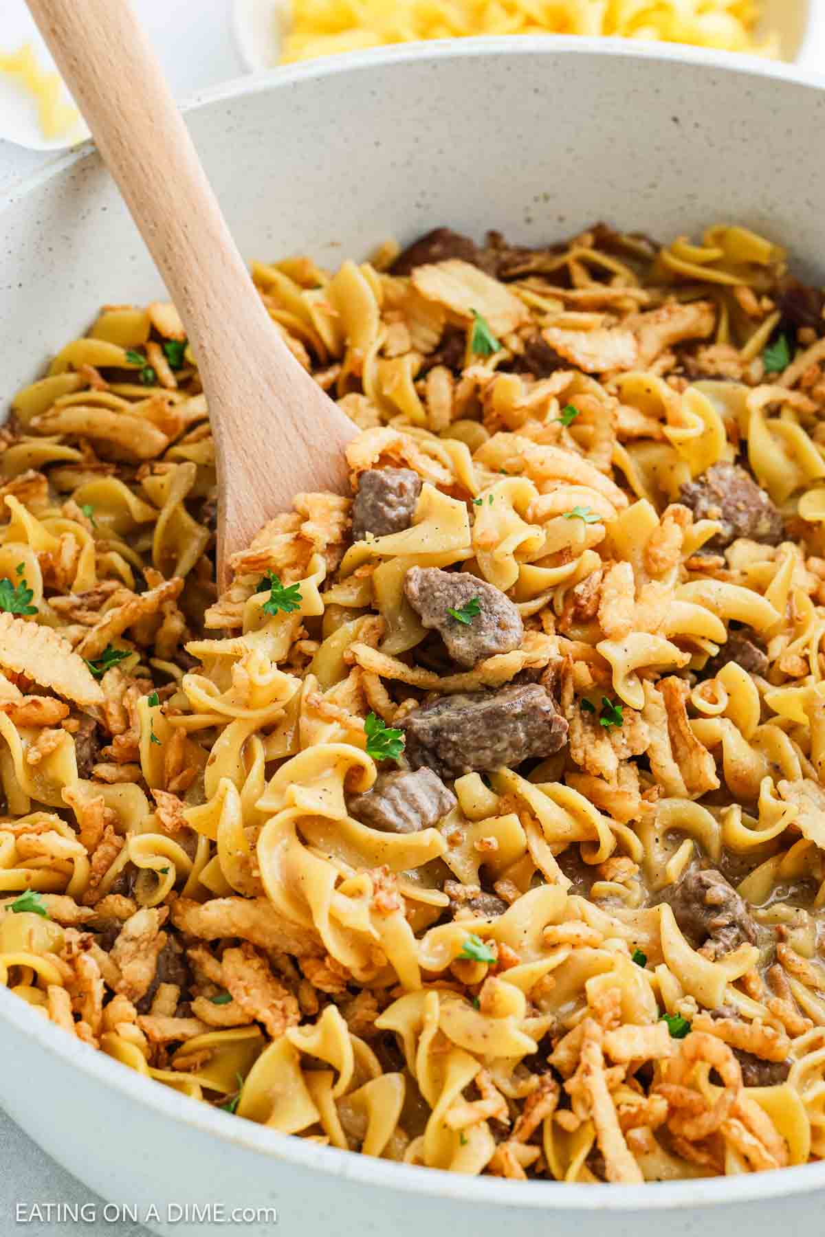 A close-up of a one pot dinner filled with French Onion Beef and Noodles stroganoff, featuring tender beef chunks, egg noodles, and parsley garnish, being stirred with a wooden spoon.