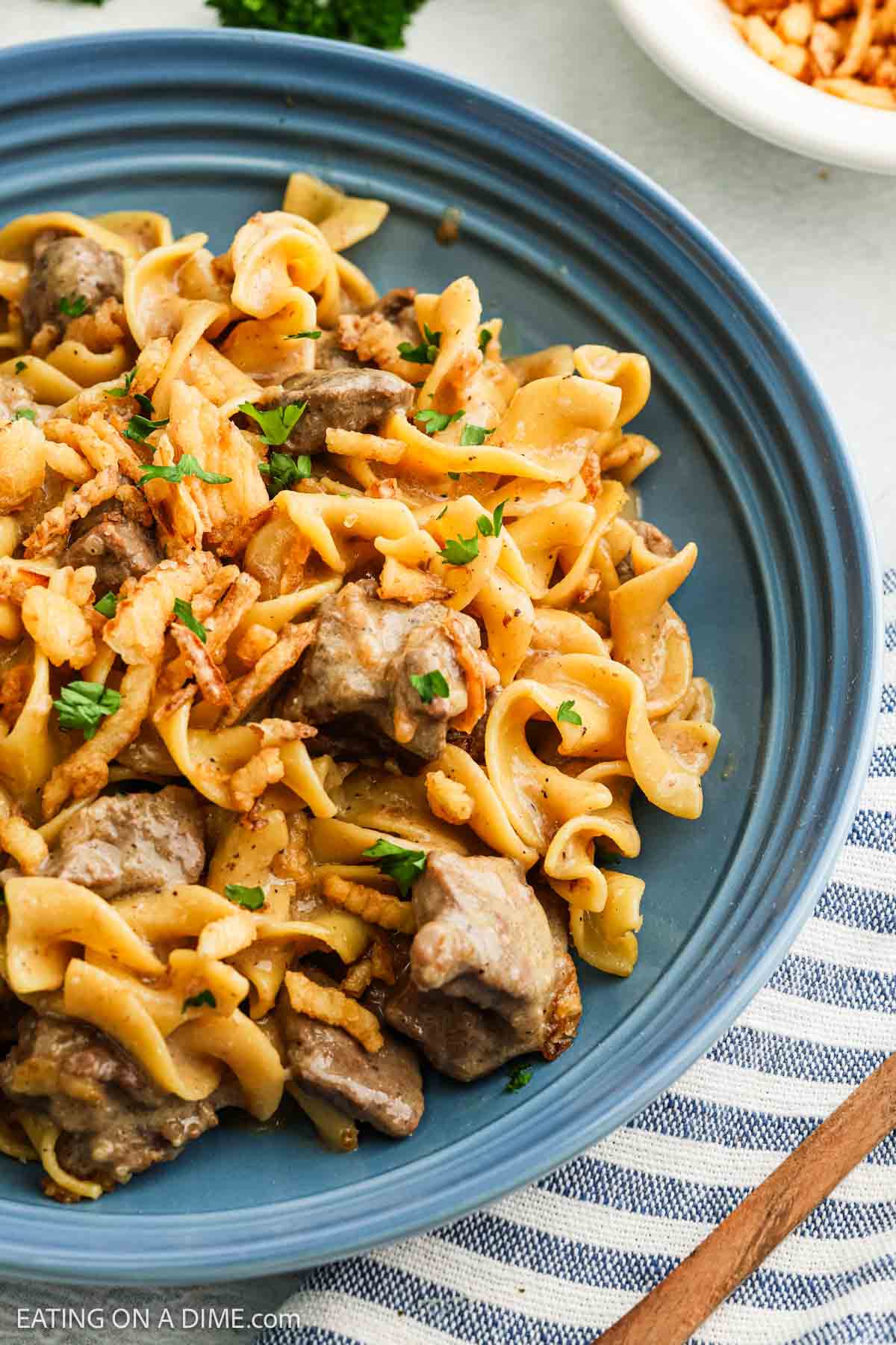 A blue plate filled with French Onion Beef stroganoff, featuring egg noodles, tender beef pieces in a creamy sauce, topped with crispy fried onions and chopped parsley. A striped cloth napkin is partially visible underneath the plate.